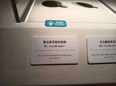 博物馆展柜中陈列着两件古代玉器，左侧为明代（公元1368-1644年）的青玉单耳乳钉纹杯，右侧为清代（公元1644-1911年）的白玉梅花单耳杯。两件玉器置于白色展台上，下方配有中英文说明牌，展柜边缘有蓝色的“036 语音导览”标识。整体画面清晰展示了文物的展陈状态，体现了博物馆对古代玉器的收藏与展示。
