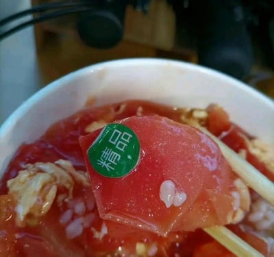 这是一碗西红柿鸡蛋盖饭，其中一片西红柿果肉上贴着一个绿色的“精品”圆形标签，标签和西红柿一起被煮在菜里，看起来十分滑稽，像是在调侃这个西红柿被标注为“精品”，却直接连标签一起被下锅烹饪，属于生活中粗心失误引发的搞笑场景。