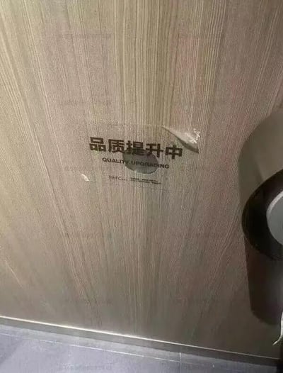 这张图拍摄了贴在木纹墙面上的“品质提升中”标识贴纸，贴纸的右上角已经翘起破损，和标识上传递的“品质提升”理念形成鲜明反差，带有讽刺意味——本该体现品质提升的标识自身都出现了损坏，显得形式大于内容，十分滑稽。
