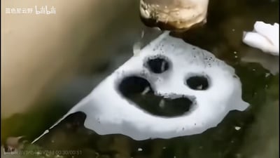这是B站趣味视频的截图，画面中水面上漂浮着一块白色的冰，冰面被人为凿出两个圆形孔洞作为「眼睛」，一个大孔洞作为「嘴巴」，形成了拟人化的笑脸造型。上方有容器正在往冰上倾倒液体，液体从冰的孔洞中流下，让这个笑脸看起来像是在「流口水」，充满了趣味性和治愈感，是利用日常物品制造的创意趣味场景。