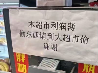 这是一张实拍的超市收银区域照片，画面主体是贴在收银设备上的白色纸张告示，告示上用黑色字体打印了内容：“本超市利润薄 偷东西请到大超市偷 谢谢”，以奇葩的反讽语气警示偷窃行为；告示下方露出了部分红底黄字的水果宣传字样，背景隐约可见超市内的货架和商品，整体内容诙谐反常，引人发笑。
