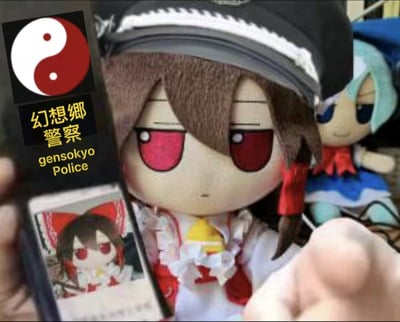 图片核心为一只红瞳卡通plush娃娃，棕色头发搭配黑白相间帽子，身着红白花边服饰，表情严肃。前景中有人手持手机对准娃娃，手机屏幕显示含阴阳符号的“幻想乡警察”（gensokyo Police）字样，下方附带该娃娃的缩略图。背景可见另一只蓝发蓝裙plush娃娃，面带微笑。整体场景呈现东方Project同人创作的趣味互动，红瞳娃娃疑似系列角色蕾米莉亚·斯卡雷特，背景蓝发娃娃推测为琪露诺。
