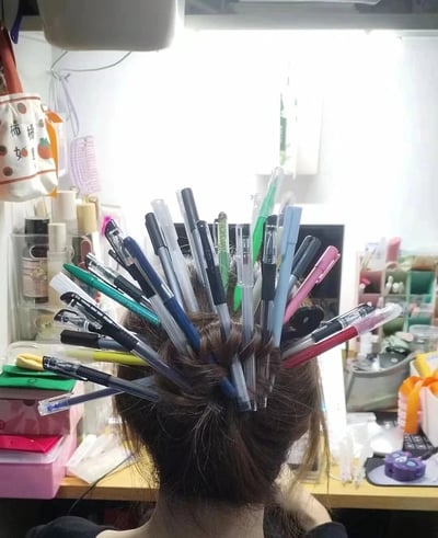 这是一张室内原始拍摄的静态图，画面主体是一位背对镜头的真实人物，她将头发盘起，在盘好的头发上插满了二十余支各色各样的中性笔，使头顶看起来像一个“移动笔筒”，充满趣味创意。背景是杂乱的个人桌面，摆放着各类美妆收纳盒、文具、电脑设备等，场景大概率是个人工位或居家书桌，这个造型像是职场打工人的搞笑整活，调侃日常工作中频繁需要使用笔的场景，充满生活化的幽默感。