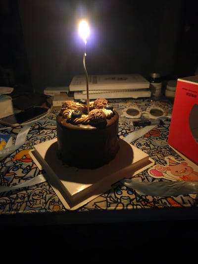一张温馨的生日场景照片，画面中央是一个小型巧克力生日蛋糕，顶部插着一根点燃的蜡烛，火焰在昏暗环境中发出明亮的光芒。蛋糕表面装饰有巧克力酱、奶油花纹和几颗新鲜浆果（蓝莓和覆盆子）。蛋糕放置在白色方形蛋糕托上，下方是一张印有卡通人物图案的彩色桌布。背景中可见几本书籍、一部手机和一个红色包装盒，整体氛围温馨而私密，展现了简单而美好的生日庆祝场景。