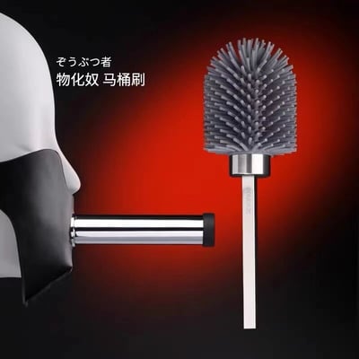 图片左侧是一个戴着黑色口塞的白色人体模型头部，口塞中插着一根金属杆；右侧是一个灰色刷毛的马桶刷，刷毛呈放射状。背景为红黑渐变，左上角有日文“ぞうぶつ者”和中文“物化奴 马桶刷”的文字组合。整体通过将人体模型与马桶刷并置，形成一种荒诞的视觉对比，可能在通过谐音或语义双关制造黑色幽默效果。