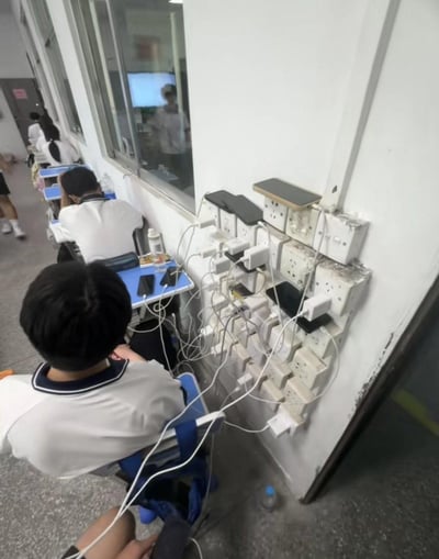 这是一间中学教室的真实场景，墙面上密集安装了大量插座与插线板，插座上插满充电器，多台手机正在充电，电线缠绕显得杂乱。教室内多名身着统一白色校服的学生，有的坐在座位上，有的在教室走动，看起来是利用课间或自习时间，集中在墙面插座区域为电子设备补充电量，墙面插座周边有明显的使用痕迹。