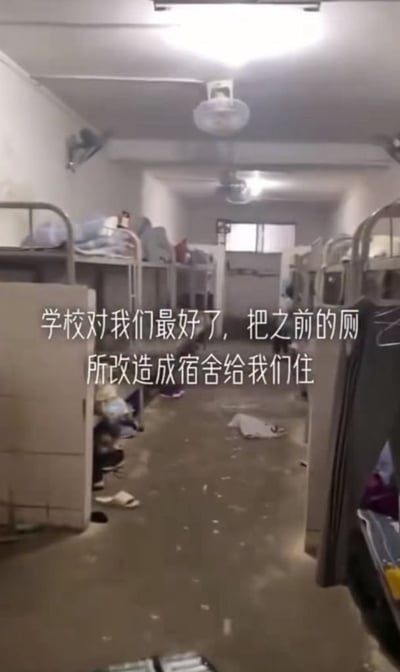 这是一张竖屏实拍图，展示了一个狭长的改造宿舍空间：房间两侧排列着多组上下铺铁架床，地面是粗糙的水泥地，散落着拖鞋、纸张等杂物，整体环境简陋杂乱。图中叠加了白色文字，以反讽的语气吐槽学校将原本的厕所改造为学生宿舍供学生居住。