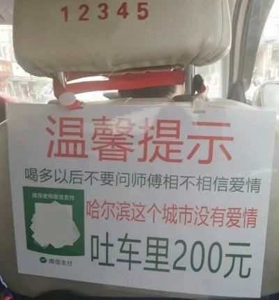 这是一张原始拍摄的静态单图，属于图文混排的顶配底文图，拍摄场景为出租车后座，画面主体是悬挂在前排座椅头枕下方的白色提示牌。提示牌顶部用醒目的红色大字标注“温馨提示”，下方依次排列三行黑色与红色混合的文字：第一行黑色文字“喝多以后不要问师傅相不相信爱情”，第二行红色文字“哈尔滨这个城市没有爱情”，第三行黑色加粗文字“吐车里200元”；提示牌左下角有绿色区域，内有被遮挡的图案，旁边配有黑色文字“推荐使用微信支付”，下方还有微信支付的标识文字。整个提示牌用幽默调侃的语气，先以网络流行的爱情玩笑话拉近与乘客的距离，最终点明乘车的核心规定，既有趣又清晰地提醒醉酒乘客注意行为，避免因呕吐弄脏车辆而产生额外费用，风格接地气且充满趣味，没有双关或谐音梗。