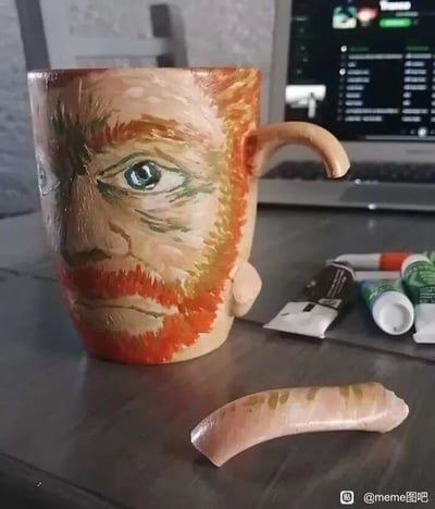 这张图片展示了一个经过创意改造的杯子，杯身手绘了著名后印象派画家梵高的肖像，肖像还原了梵高标志性的红棕色胡须和凝重的神情。原本的杯把手断裂，断裂下来的部分被制作成类似人类耳朵的形态，放置在杯子前方的桌面上，巧妙呼应了梵高曾割下自己耳朵的著名轶事。杯子的右侧摆放着几支颜料管，暗示了这个创意是通过手绘改造完成的，背景中还能看到一台打开的笔记本电脑，整体场景呈现出一种充满趣味和艺术梗的手工创作氛围。