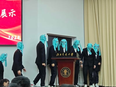 这是2024年北京交通大学某展示活动的现场实拍画面，一群身着正式黑色西装的学生，戴着自制的魔性浅蓝色初音未来样式头套，在印有北京交通大学校名的讲台附近活动，有的聚集在讲台旁、有的侧身走动，严肃的正装与滑稽的头套形成强烈反差，营造出十足的搞笑氛围，画面中没有双关、谐音梗或暗示内容。