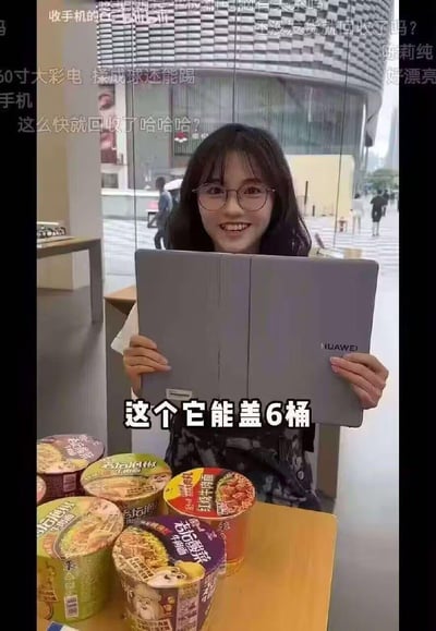 一位戴眼镜的女生在室内门店中，双手举着一台中间弯折的华为大尺寸二合一平板/笔记本，面前的桌子上整齐摆放着6桶不同口味的杯装泡面。图中配文“这个它能盖6桶”，用调侃的方式玩“电子产品当泡面盖”的经典梗，吐槽大尺寸电子产品的另类“实用”用途。画面上方的弹幕还有各种搞笑评论，比如“60寸大彩电 揉成球还能踢”“收手机的 还没发货就回收了吗？”，整体氛围轻松欢乐，用夸张的演示制造笑点，调侃了网友们常说的“买前生产力，买后泡面盖”的电子产品使用现状。