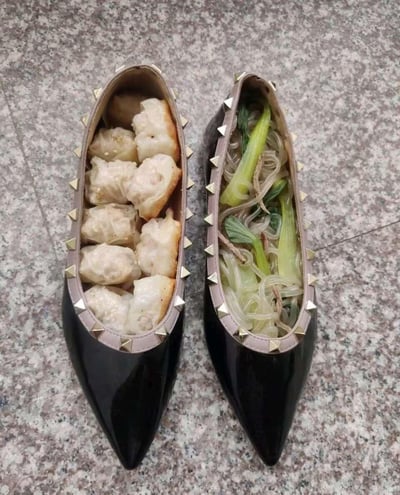 这是一张充满恶搞创意的实拍图：一双带有金属铆钉装饰的黑色尖头漆皮女鞋，被当作食物容器使用。左侧鞋子里盛放着煎制过的肉馅云吞（或煎饺），表皮带有煎制后的焦黄色泽；右侧鞋子里装着粉丝青菜汤，透明粉丝搭配翠绿青菜，视觉冲击力拉满。用日常穿的鞋子来盛装食物，这种反常规的摆盘方式充满猎奇感，主打一个脑洞大开的搞笑创意，没有双关或谐音梗，纯靠意想不到的搭配制造趣味。