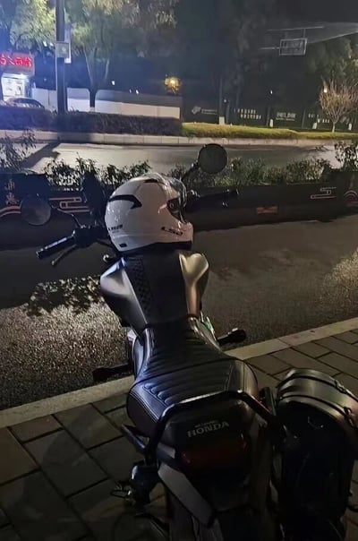 夜晚的街道旁，一辆黑色本田摩托车停放在砖砌人行道上，车把上放置着一个白色头盔。背景可见路灯、树木、篮球架及带有灯光招牌的建筑物，地面湿润，可能刚下过雨，反射着周围的灯光，营造出宁静的城市夜景氛围。