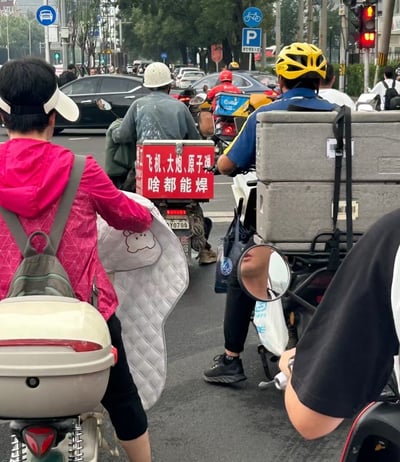 这张图片拍摄于城市道路路口，许多骑电动车的人在等待红灯。画面中心一位骑着电动车的男子，后座上挂着一个红底白字的广告牌，内容是“飞机、大炮、原子弹，啥都能焊”。这句广告词极度夸张、幽默风趣，显然不是真的意思，而是用来表达这个师傅焊什么都行，有种“无所不能”的调侃感。广告语本身采用了网络段子的夸张表达方式，具有无厘头和戏谑色彩，令人会心一笑。这类表达方式在民间广告和网络平台上十分流行。图中整体氛围轻松搞笑，体现了中国街头独有的幽默文化。