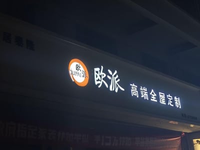 这是一张在夜晚拍摄的线下门店招牌照片，画面主体是带有灯光的「欧派 高端全屋定制」标识，左侧配有橙色圆形的欧派品牌LOGO，LOGO内包含“欧派”与“OUPAI”字样。招牌左上角还有“居泰隆”字样，右下角标注了联系电话“8723558”，招牌下方有模糊的红色促销横幅，整体环境偏暗，招牌的灯光突出了品牌的业务信息，展示了欧派家居线下门店的夜间外观。