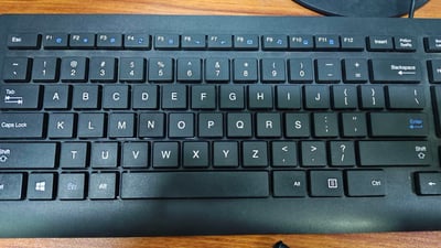 这是一张实拍的黑色台式电脑键盘照片，展示了完整的主键盘区以及上方的F1-F12功能键区，按键采用美式QWERTY布局，包含字母键、数字符号键、常用控制键（如Backspace、Enter、Shift、Ctrl等），键盘放置在浅棕色木质桌面上，是日常办公、电脑操作的通用输入设备，外观简洁实用。