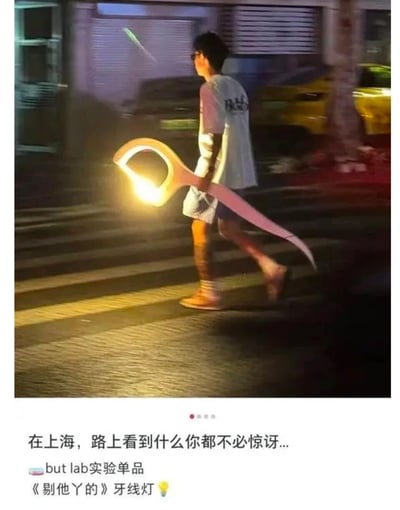 这是一张夜晚的上海街头实拍图，画面中一名戴眼镜的男生正行走在斑马线上，他手中拿着一个造型为巨型牙线的实验灯具——but lab的《剔他丫的》牙线灯，灯具的手柄部分散发着暖黄色的光，整体造型夸张又充满创意。图下方的配文“在上海，路上看到什么你都不必惊讶...”，既点出了拍摄地点，也凸显出这件创意单品的新奇感，让日常的街头场景变得趣味十足。