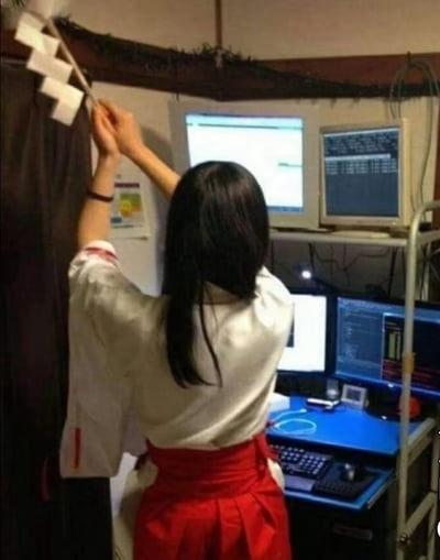一位身着传统服饰的女性站在多台电脑显示器前，双手举着一个带有锯齿状图案的白色纸制品。她的上半身穿着白色上衣，下半身搭配红色裙子，长发披肩。背景中可以看到至少四台电脑显示器，显示着不同的内容，暗示这是一个技术工作环境。传统服饰与现代科技设备形成鲜明对比，营造出一种文化交融的视觉效果。