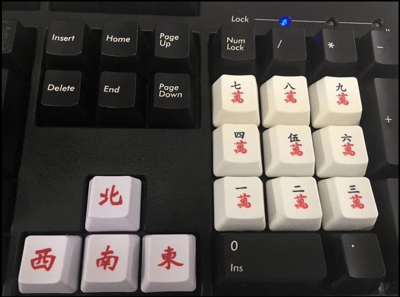 一张黑色机械键盘的特写照片，键盘右侧数字小键盘区域及部分功能键被创意替换成白色麻将牌键帽。其中数字键区域整齐排列着印有红色文字的“一万”至“九万”麻将牌键帽，呈3x3矩阵分布；左下角另有一颗“北”字麻将牌键帽。其余按键仍为原装黑色键帽，印有白色标准键盘字符（如Insert、Home、Delete等），右上角Num Lock指示灯呈蓝色亮起。整体展现了将传统麻将元素与现代电脑外设结合的趣味DIY改装，充满生活气息与创意巧思。