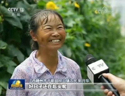 图片为CCTV 13新闻频道的采访截图，画面中是江西赣州市于都县梓山镇农民郭月华，她穿着紫色碎花衬衫，面带灿烂笑容，正在接受记者采访。背景是郁郁葱葱的绿色植物和黄色花朵，前方有带有“CCTV”标识的麦克风。屏幕底部字幕显示“江西赣州市于都县梓山镇农民郭月华 好日子还在后头呢”，传递出对未来生活的美好期盼。