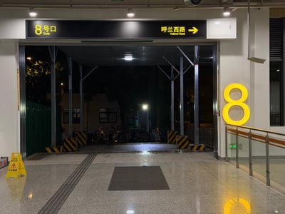 这是一张夜间拍摄的地铁站8号出口实拍图，出口上方的黑色标识牌左侧标注黄色字样「8号口 Gate」，右侧标注「呼兰西路 Hulanxi Road」及右向箭头，出口右侧墙面设有醒目的亮黄色立体数字「8」标识，出口外环境昏暗，仅能隐约看到停放的车辆和远处灯光，地面铺设有灰色地垫，左下角放置有黄色「小心地滑」警示牌。该场景恰好对应恐怖游戏《8番出口》的名称，昏暗未知的出口环境也和游戏中诡异的通道设定相似，具有玩梗属性。