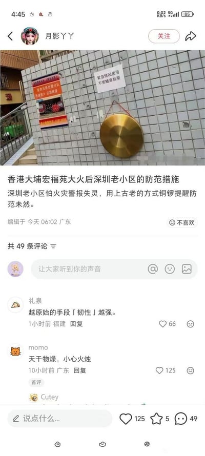 社交媒体帖子截图，内容为深圳某老小区在香港大埔宏福苑大火后采取的消防安全防范措施。图片主体是小区白色瓷砖墙壁，上面挂着一面金色铜钲（铜锣），旁边贴有一张白色告示，写着“紧急情况使用 不要随意玩耍”。帖子配文说明因小区火警报警器可能失灵，故采用传统铜钲这种古老方式提醒居民防范火灾。截图下方显示有49条评论，部分评论内容为“越原始的手段[韧性]越强”“天干物燥，小心火烛”等。