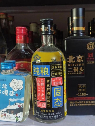 这张图片展示了货架上陈列的多款国产白酒，前景有三款清晰可见的酒品：左侧是一款蓝色包装的小酒，瓶身印有“北嘎达”字样和东北雪屋图案，标注酒精度42%vol，净含量125ml；中间是杏花村酒业的纯粮固态白酒，透明瓶身搭配黄黑配色标签，显眼标注“国标优级 大曲清香”，酒精度42%vol，净含量475ml，还标注有“二十年老酒勾调”以及总酯、总酸的含量标准；右侧是北京二锅头（永丰版），黑色瓶身搭配金色装饰，标注“永丰传奇 -1163-”，酒精度42%vol，净含量480ml。背景中还有其他模糊的酒瓶，整体场景像是在酒品售卖的货架或者陈列柜中，展示了不同品牌、包装和规格的清香型白酒。