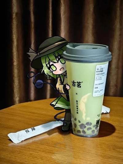 这是一张创意合成的趣味图片，将《东方Project》中的Q版角色古明地恋通过P图方式，合成到古茗抹茶布蕾奶茶的实拍场景中。角色的绿发绿裙与抹茶奶茶的淡绿色调完美呼应，做出双手环抱奶茶杯的亲昵动作，仿佛正和喜爱的奶茶贴贴，旁边还摆放着一根蜜雪冰城的糖包，整体画面软萌治愈，是ACG爱好者将二次元角色与三次元美食结合的趣味二次创作，没有双关或谐音梗。