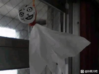 这是一张原始拍摄的单图，展示了一个用户自制的搞怪晴天娃娃：娃娃的身体由白色布料制成，头部是圆形白色物体，上面用黑色马克笔手绘了暴走漫画风格的搞怪脸——双眼不对称斜吊，嘴巴歪扭着露出诡异的笑容，娃娃被红色绳子系住悬挂起来，背景是室内的墙面与门框。原本晴天娃娃是带有温柔祈福意味的传统小物，这张图将其改成了反差感拉满的搞怪形象，整体风格滑稽搞笑，没有双关、谐音梗等特殊暗示内容。