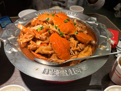 这是一张线下餐厅用餐时拍摄的实拍美食图，画面主体是胖哥俩品牌的肉蟹煲，金属制的大份餐锅边缘刻有品牌名称“胖哥俩”字样，锅内盛放着烹制完成的肉蟹煲：表层铺有多只色泽橙红的熟梭子蟹，蟹下搭配了炖煮至软嫩入味的鸡爪，所有食材浸在红棕色的浓郁酱汁中，表面撒有碎葱花点缀提色，锅边摆放着金属汤勺，周围桌面还能看到盛放的白米饭、水杯、手机等用餐相关物品，整体呈现出日常外出就餐的烟火气场景。