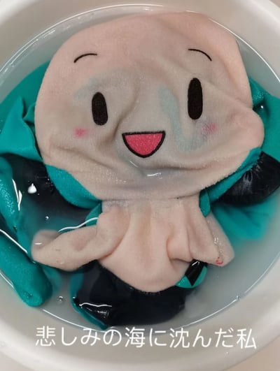 这是一张实拍的初音未来主题毛绒玩偶图片，玩偶被泡在水盆的水中，布料浸湿后皱缩，脸部还出现晕开的蓝色痕迹，看起来像是在“哭泣”。图下方配有日文文字“悲しみの海に沈んだ私”，翻译为“沉入悲伤之海的我”，用日常清洗玩偶的场景玩梗，将玩偶浸湿的状态拟人化，营造出悲伤又带点搞笑的反差感，用来表达低落、emo的情绪。
没有双关或谐音梗，主要是通过场景与文字的结合，把普通的洗衣场景转化为“沉浸悲伤”的拟人化情绪表达。
