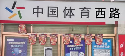 图片展示了一处店铺的招牌区域，上方为白色底红色边框的招牌，左侧有彩色几何图形标志，右侧是黑色大字“中国体育西路”。招牌下方的玻璃窗上悬挂着多个红色包装的宣传物料，玻璃窗内侧贴有蓝色海报，内容包括“测测我的购彩风格 够不够理性？”和“问题线索征集公告”等文字，整体看起来像是体育彩票或相关服务的店铺。