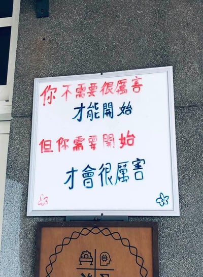 这是一张实拍的墙面告示照片，灰色粗糙质感的外墙上挂着两块告示板，靠上的白色边框白板用红蓝两色手写了四行文字，红色字为「你不需要很厉害」「但你需要开始」，蓝色字为「才能开始」「才会很厉害」，白板左下角和右下角还分别画了粉色和蓝色的简笔画小花作为装饰，白板下方是一块棕色边框的木质告示牌，边缘有波浪形装饰线条，牌面带有简单的图标图案。整体内容传递出无需等到足够优秀才开始行动，行动起来才能变优秀的正向鼓励导向。