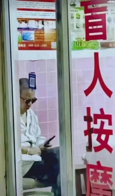 这是一张在盲人按摩店门外拍摄的静态照片，透过玻璃门可以看到店内一位戴着墨镜的男士正坐着低头看手机。由于玻璃门的位置巧合，玻璃上的一个付款二维码刚好和男士的头顶位置重合，从门外的视角看去，就像是这个二维码直接长在了男士的头顶上，旁边的玻璃上还印有红色的“盲人按摩”字样，这个意外的视觉错位形成了十分搞笑的效果，让人忍俊不禁。