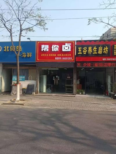 这是一张户外拍摄的临街商铺照片，画面呈现了三家相邻的本地美食店铺：左侧是蓝色招牌的「北伴岛·海鲜」，中间是红色招牌的卤味店「帮你卤」，标注了卤货品类、招牌菜品及微信联系方式；右侧是红黑配色的「五谷养生磨坊」，带有联系电话，门口电子屏滚动显示「发 少放 烟花爆竹」的提示语。店铺前的人行道上有一棵行道树，整体是平实的日常街边商业场景，没有谐音梗、双关或暗示类内容。