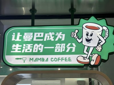 图片展示了曼巴咖啡(MAMBA COFFEE)的店铺招牌。招牌主体为绿色背景，左侧有白色中文标语“让曼巴成为生活的一部分”，下方标注英文品牌名“MAMBA COFFEE”并配有一个咖啡滴图案。右侧是一个拟人化的卡通咖啡杯形象：白色杯身，内盛红色液体，面带微笑，左手叉腰，右手竖起大拇指，脚穿红白相间的鞋子，整体设计活泼亲切。招牌通过绿色支架悬挂于天花板，背景可见店铺内部的金属结构和部分装修。