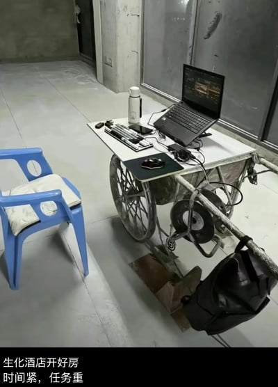 在未装修完成的空旷房间（疑似工地或待装修房屋）里，网友用工地手推车改造成临时办公桌，上面摆放着笔记本电脑、外接键盘、鼠标、保温杯等设备，旁边搭配一把蓝色塑料椅加坐垫，打造出一个硬核的办公/游戏工位。底部配文「生化酒店开好房 时间紧，任务重」，其中「生化酒店」是游戏《穿越火线》中的经典地图，结合简陋的场景玩梗，形成强烈反差，调侃在艰苦环境下依然赶工或打游戏的状态，搞笑又充满无奈感。