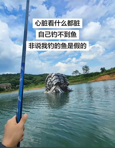 这是一张经过PS合成的搞笑玩梗图，采用第一人称视角，画面左下角是一只手握着蓝色钓鱼竿，身处开阔的户外水库场景，水库周围是绿植覆盖的山坡和蓝天白云。水库水面中突兀地露出了特摄作品《迪迦奥特曼》中的最终BOSS加坦杰厄的半身，画面上方配有三行白底黑字的文字：“心脏看什么都脏，自己钓不到鱼，非说我钓的鱼是假的”，用钓到根本不可能存在的巨型怪兽这种极度夸张的内容，反讽那些自身能力不足做不到某件事，就恶意质疑他人成果、喜欢酸别人的群体。
