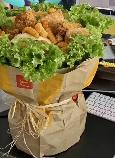 这是一款极具创意的麦当劳美食花束，用麦当劳的外卖包装纸将生菜、炸鸡、薯条等食物包裹成花束造型，并用麻绳捆绑固定。花束以生菜作为装饰衬托，中间填满了香酥的油炸小食，放置在办公桌面，背景可见键盘，把浪漫的花束形式和美味的快餐结合在一起，既拥有花束的仪式感，又能享用美食，是很有趣的创意礼物，没有双关或谐音梗。