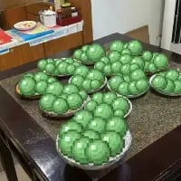 这是一张真实拍摄的生活照片，深色桌面上摆放着多盘色泽翠绿的青团，每一盘都被青团满满当当填满，数量十分可观。背景中能看到室内的桌椅和一些杂物，整体场景给人一种即将享用或分发传统美食的温馨丰盛感，青团作为江南地区的传统寒食节美食，通常在清明前后食用。