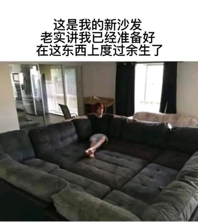 这是一张文字加注的实拍生活图，画面展示了一个室内客厅场景，核心是一套尺寸远超常规的L型组合沙发，沙发由深灰色和浅灰色的模块拼接而成，占据了画面的绝大部分空间，显得十分宽大。沙发中间坐着一位红头发的人，他姿态慵懒放松，一只腿弯曲，一只脚搭在另一条腿上，手臂搭在沙发靠背上。图片上方配有醒目的中文文字：“这是我的新沙发 老实讲我已经准备好在这东西上度过余生了”，直白地表达了沙发主人对这套巨型沙发的喜爱，传递出一种对舒适、慵懒居家生活的向往，让人感受到这个沙发带来的极致舒适感。