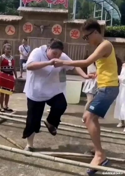 这是一处民俗类景区的户外互动体验场景，背景建筑上的文字显示为“人 洞 欢 迎”，顶部被遮挡的红色大字应该是“仙人洞”，推测是名为仙人洞的景区。画面中一位穿白色上衣的女士和一位穿黄色背心的男士正在尝试跳竹竿舞，女士动作慌乱明显没跟上节奏，男士伸手拉着她试图帮她跟上节拍，旁边还有身着民族服饰的工作人员和其他游客围观，整个场景充满了欢乐搞笑的氛围，是典型的游客游玩时的趣味瞬间。