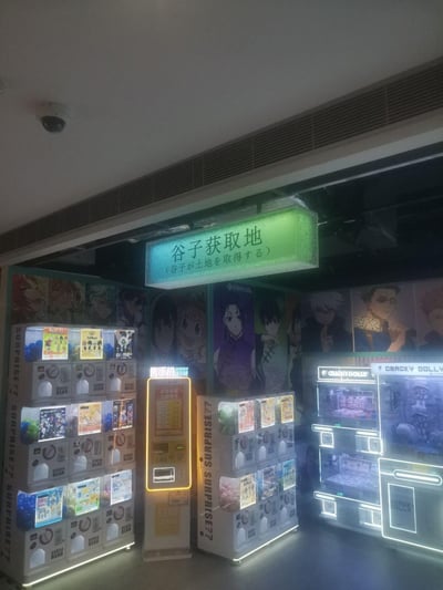 这是一处线下的二次元周边自动售卖区域，上方悬挂着带有中日双语的发光招牌“谷子获取地（谷子が土地を取得する）”，区域内摆放着多台标有“SURPRISE??”“CRACKY DOLLY”字样的扭蛋机，以及一台售币机，扭蛋机内展示着不同动漫IP的周边产品。背景墙印有《咒术回战》等热门动漫的角色海报，整体场景面向二次元爱好者，“谷子”是二次元圈对“goods（周边）”的谐音梗，用来指代各类动漫衍生周边产品，营造出轻松的二次元消费氛围。