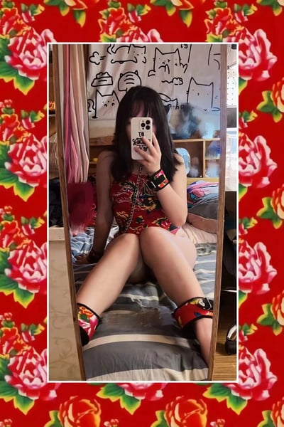 一位穿着红色碎花连体装的女生在卧室镜子前自拍，手机挡住了面部。她搭配了同系列的红色碎花脚链和手链，背景墙上有可爱的猫咪涂鸦。整个照片被喜庆的红色花卉边框包围，营造出浓厚的中国风氛围。