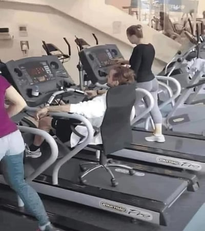 这是健身房内的搞笑场景：一位男士将办公椅放置在跑步机上，整个人躺靠在椅子上，摆出类似使用健身器材的姿势，实则是在摸鱼休息。旁边的健身者都在认真使用椭圆机、跑步机锻炼，形成了强烈的行为反差，充满了搞笑的氛围，完美诠释了健身房里的极致摸鱼操作。