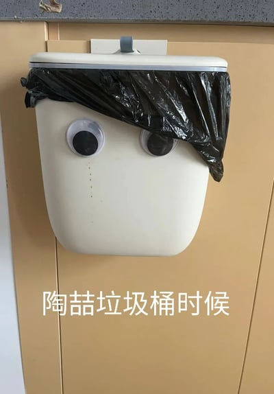 这是一张拍摄日常厨房挂式垃圾桶的趣味照片，米白色挂式垃圾桶被贴上活动眼睛，桶口的黑色垃圾袋被摆放成类似头发的造型，桶身还有几处点状痕迹类似泪痕，将垃圾桶拟人化为一个委屈的小角色。图片底部配有文字“陶喆垃圾桶时候”，利用“陶喆”与“掏这”的谐音制造玩梗效果，实际意思是“掏这个垃圾桶的时候”，让原本普通的生活物品充满趣味性，是网友对日常物品的创意改造。