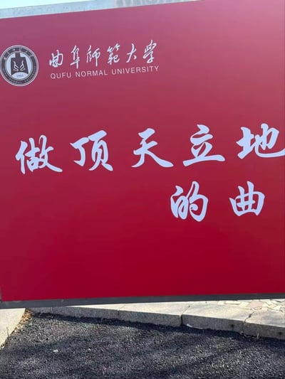 图片展示了曲阜师范大学的一块红色大型标语牌。牌面顶部左侧有学校校徽，右侧为'曲阜师范大学'的中文校名及英文翻译'QUFU NORMAL UNIVERSITY'。牌面中央是白色书法字体书写的标语'做顶天立地的曲'。背景为纯红色，底部可见部分地面和石质基座。