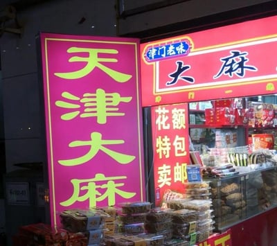 这是一张线下天津特色小吃店铺的实拍照片，店铺主营天津大麻花这类传统点心。画面左侧是亮粉色底配黄绿色大字的竖版招牌，写着“天津大麻”；右侧红色招牌带有“津门老味”标识，同样标注“大麻”，下方还有排版特殊的“花额特卖 包邮”字样（原文字排版为“花额 特包 卖邮”）。店铺柜台前堆叠着多盒包装好的麻花等点心，玻璃柜台内也陈列着各类麻花产品。此处的“天津大麻”是“天津大麻花”的简称，因省略了“花”字造成歧义，容易让观者产生误解。