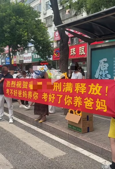 这张图是国内高考结束后考点外的实拍视频截图，画面中几位家长站在人行道上共同举着一条红底黄字的横幅，横幅将高考结束调侃为“刑满释放”，还写有“考不好爸妈养你 考好了你养爸妈”的内容，旁边还摆放了准备送给孩子的花束，背景可见沿街商铺、行道树和公交站台，家长用诙谐轻松的方式迎接考完的孩子，既调侃了备考阶段的高压状态，也传递出对孩子无条件的包容与爱意。