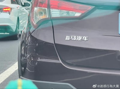 这是一张在城市道路车流中拍摄的黑色汽车尾部照片，原本的海马汽车尾标被修改为“每马汽车”，通过替换“海”与“每”的汉字制造谐音梗，充满趣味感。画面背景还能看到一辆白色的汽车，整体场景是日常道路通行的状态。
谐音梗说明：将“海马汽车”改为“每马汽车”，是利用汉字字形相近的特点玩的文字玩笑，制造出荒诞搞笑的效果。
